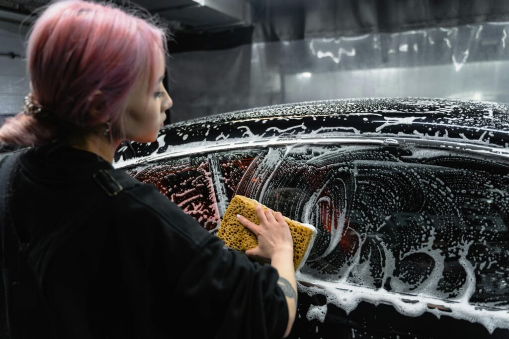 Woman washing black car with sponge, creating foamy soap suds indoors.